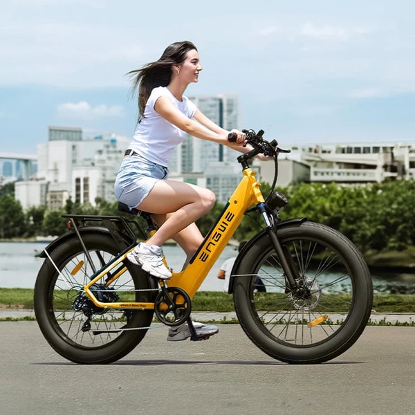 Vrouw die rijdt op een gele Engwe E26 ST elektrische fatbike langs een kade met een stadsgezicht op de achtergrond