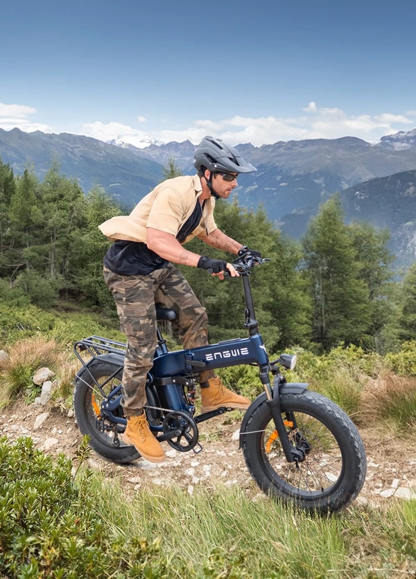 Helmeted rider in a khaki shirt and camo trousers riding a blue Engwe Engine Pro 3.0 Boost folding electric fat bike on a rocky mountain trail with pine trees and snow-capped peaks behind