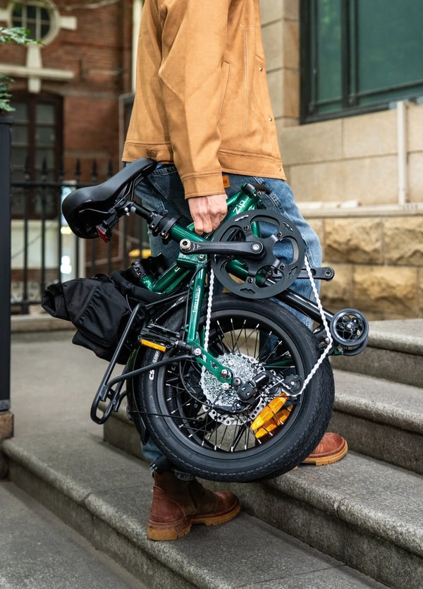 Persoon die een opgevouwen groene Engwe Zip elektrische fiets draagt over een buitentrap, met het compacte opgevouwen frame, wielen en voorste opbergtas