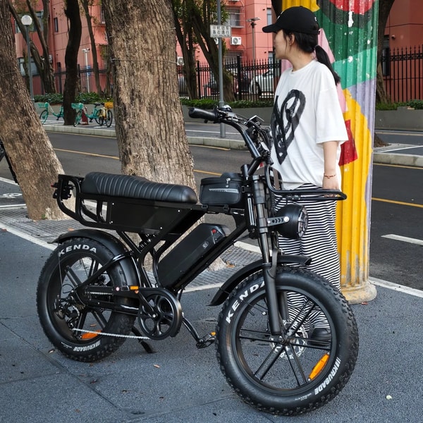 Vrouw staand naast een zwarte VOMO X90 elektrische fatbike op een stadsstraat, die de voorste bagagedrager, het lange gewatteerde zadel, de ronde koplamp en brede banden toont
