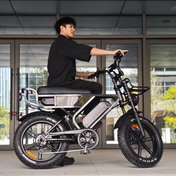 Man zittend op een blauwe VOMO X90 elektrische fatbike buiten een modern gebouw, die het lange gewatteerde zadel, voorvering, verwijderbare accu en fatbike banden toont
